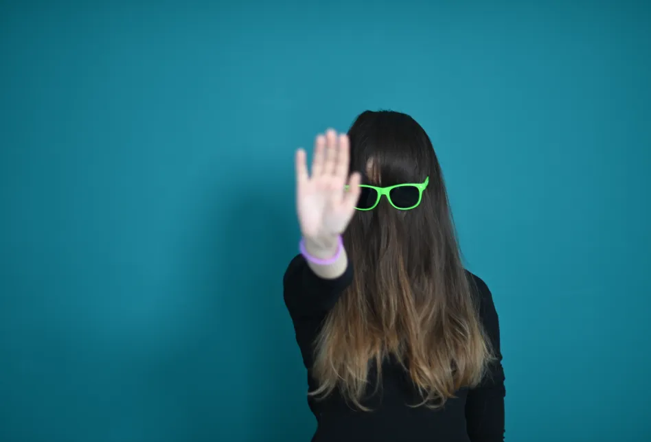 Ein Mensch macht eine abwehrende Handbewegung und hat die langen Haare so, dass sie das Gesicht verdecken. Darüber eine Sonnenbrille.